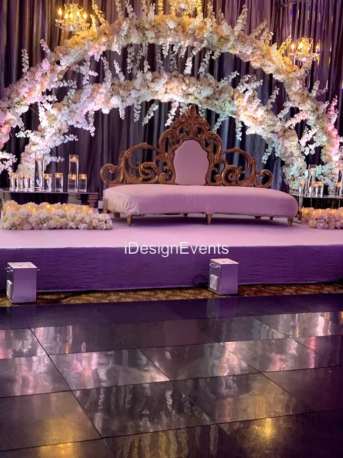 Elegant stage setup with white floral arches and a white couch on a purple carpet.
