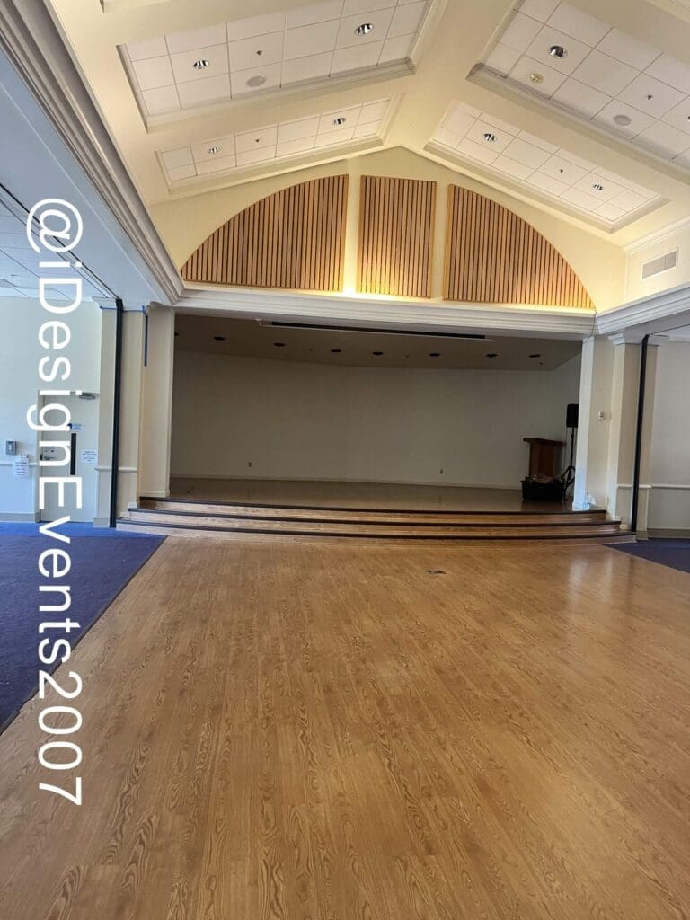Empty event hall with wooden floor and stage under arched ceiling.