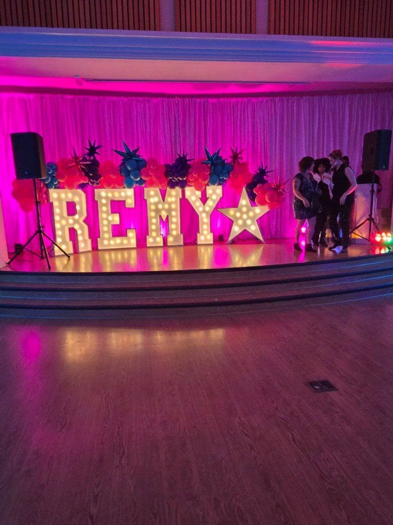 Illuminated letters spelling 'REMY' with decorative stars on a stage with pink lighting.